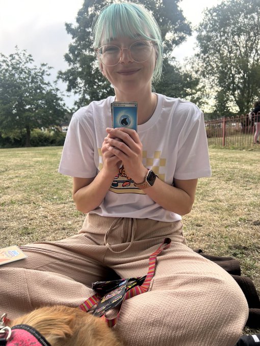 A young woman with mint green hair and round glasses sits on grass holding a phone, wearing a white t-shirt and beige pants, with a small dog on a leash beside her.