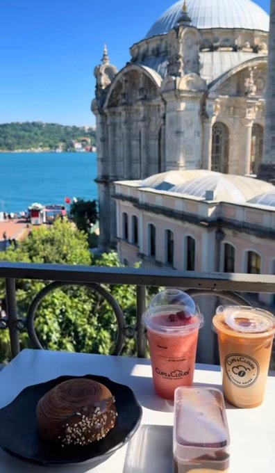 A scenic cafe balcony view featuring a pastry and iced beverages (pink raspberry and iced coffee) on a table, overlooking an ornate domed mosque and the Bosphorus strait in Istanbul.