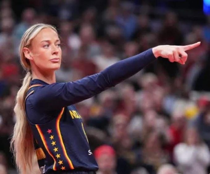 A blonde gymnast in a navy leotard with stars and the name "NOAH" visible points confidently off-camera, while the caption humorously suggests her stern "you didn't tell me" expression matches the feeling of a girlfriend discovering a hidden crypto profit.