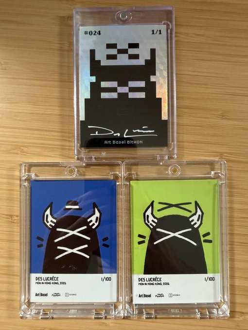Three trading cards encased in protective sleeves are displayed on a wooden surface, featuring pixel-art character designs by artist Basel BAHON. The top card is a rare 1/1 numbered #024, while the two bottom cards from the "DES LUCRECE PUSH IN FOMO FOMO 2026" series are limited to 1/100 editions—one with a blue background and one with a neon green background—showcasing the artist's signature character artwork.