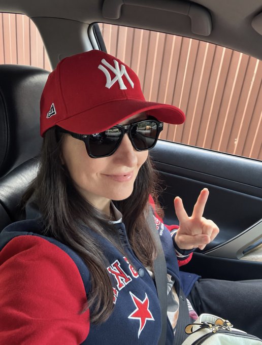 A smiling woman sits in the passenger seat of a car wearing a red NY Yankees cap, dark sunglasses, and a navy and red varsity jacket, making a peace sign with her fingers.