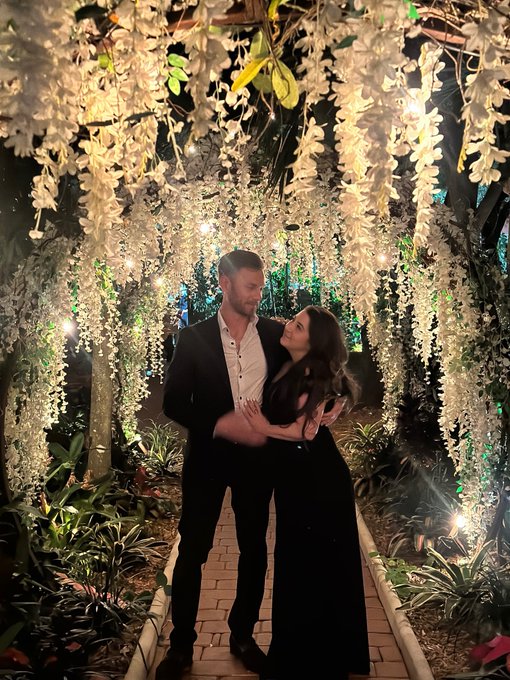 A couple embraces under a tunnel of hanging white flowers illuminated by warm string lights at night.