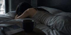 The image shows a woman lying down on her bed with her head resting on her hand, possibly enjoying some relaxation time or taking a break from her daily activities. A cup of coffee is placed next to her on the bed, suggesting that she might be having a drink while unwinding in her bedroom.