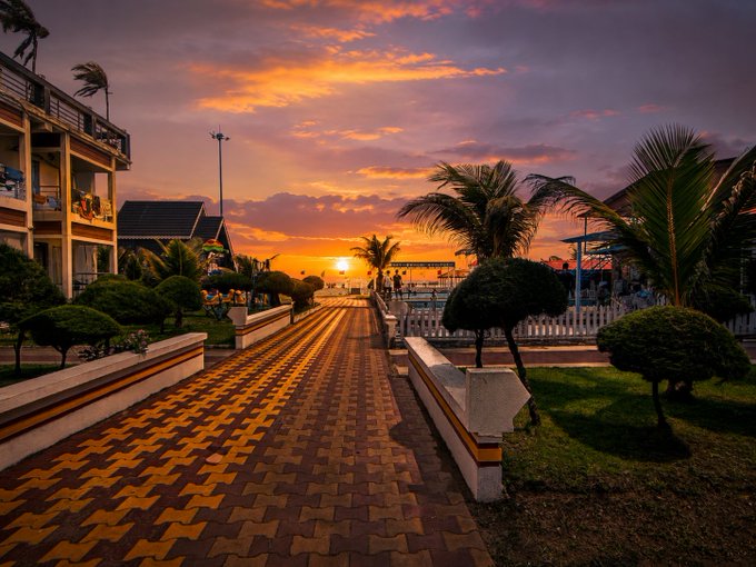 A scenic sunset photograph of a paved walkway lined with palm trees and manicured shrubs leading toward the ocean, with people walking along the path and buildings visible on either side under a vibrant orange and purple sky.
