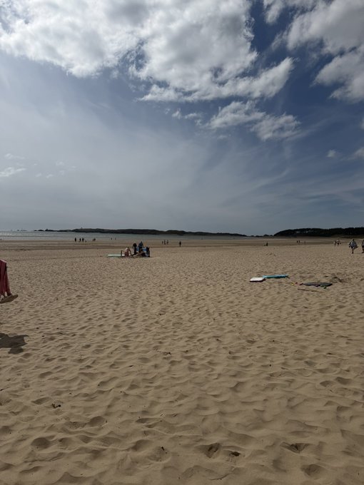A serene, expansive sandy beach stretches toward the horizon under a partly cloudy sky, with a small group of people gathered in the distance, likely enjoying a game of DDLTCG (Don't Drop The Combo, Gringo or similar beach card game). The relaxed coastal scene perfectly complements the post's caption about a new favorite hobby of playing a card game on the beach.