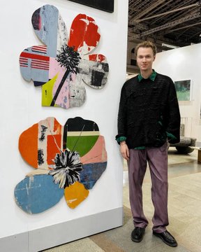 A man stands beside a colorful abstract artwork displayed on a white gallery wall, wearing a dark patterned sweater and mauve trousers.
