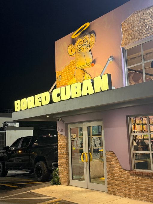 A nighttime exterior shot of a restaurant named 'BORED CUBAN' with yellow illuminated signage and a cartoon monkey mural holding a cigar and wearing a halo above the entrance.