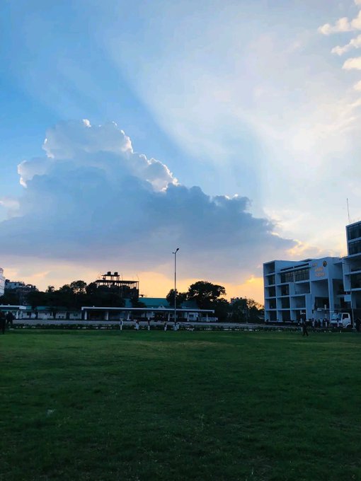 A scenic photograph of a large grassy field at sunset, with beautiful crepuscular rays beaming through clouds over campus buildings and trees.