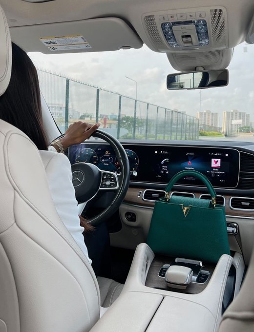 An interior shot of a car showing a woman driving, with a green Louis Vuitton handbag placed on the passenger seat.