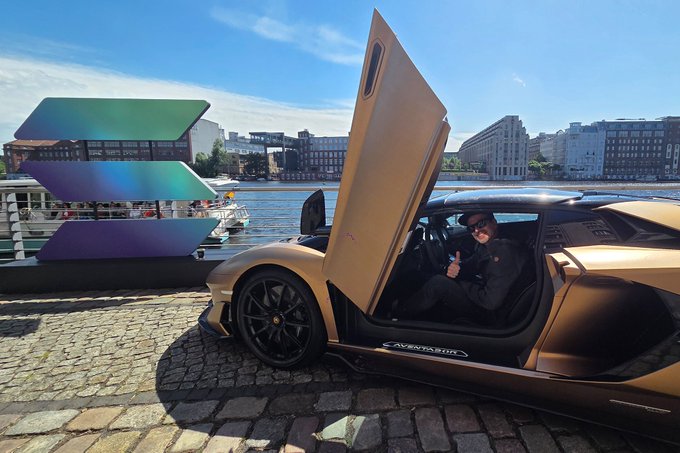 A bronze Lamborghini Aventador with its scissor doors open sits on a cobblestone street, with a person inside giving a thumbs up; the word 'AVENTADOR' is visible on the side.