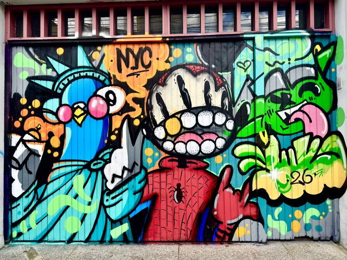 A colorful street art mural on a garage door featuring a blue bird-like character, a central figure with a large grinning mouth showing teeth, and a green creature, with 'NYC' written at the top and '126' on the right side.