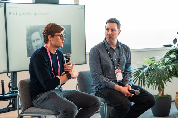 Two men sit on chairs during a panel discussion, with the speaker on the left holding a microphone while the attentive listener on the right also holds a microphone. Behind them, a screen displays "Building for the agentic web" alongside a photo of another participant. The scene captures the speaker from @p0 discussing their Index product with the questioner, as referenced in the post.