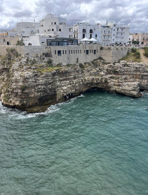 White-washed buildings of a coastal cliffside town sit above rocky limestone cliffs and turquoise Mediterranean waters under a cloudy sky.