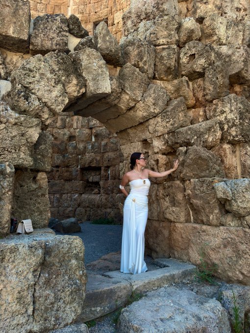 A woman in a flowing white dress and sunglasses poses inside an ancient stone structure, touching the massive weathered blocks of the wall.