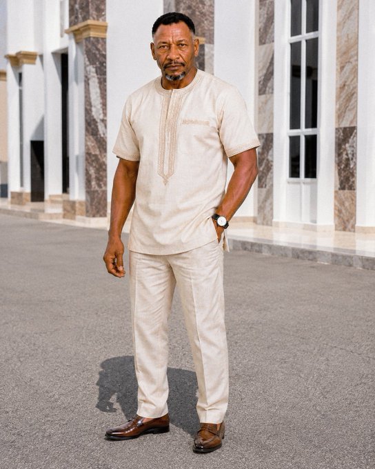 A man models an elegant cream-colored short-sleeve tunic with embroidered detailing and matching linen trousers, paired with brown loafers, standing in front of marble columns. The caption "You asked for our Sunday outfit. Here is mine." perfectly complements the polished, traditional attire styled for a refined weekend look.