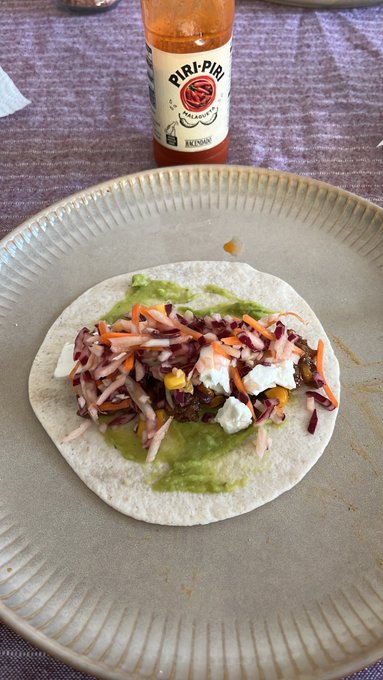An open tortilla topped with guacamole, red cabbage slaw, carrots, corn, and crumbled cheese on a beige plate, with a bottle of Piri-Piri hot sauce in the background.