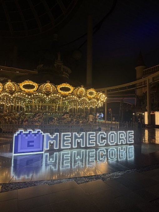 A nighttime photo of an illuminated carousel with a large neon sign reading 'MEMECORE' with a purple brick icon, reflected on the wet pavement in front of it.