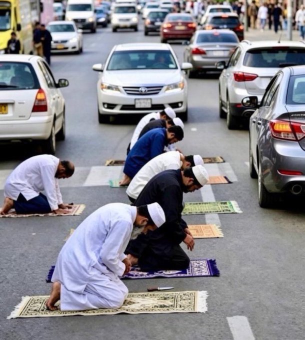 Muslim men praying on prayer rugs placed in the middle of a busy street between cars, wearing white prayer caps and traditional clothing.