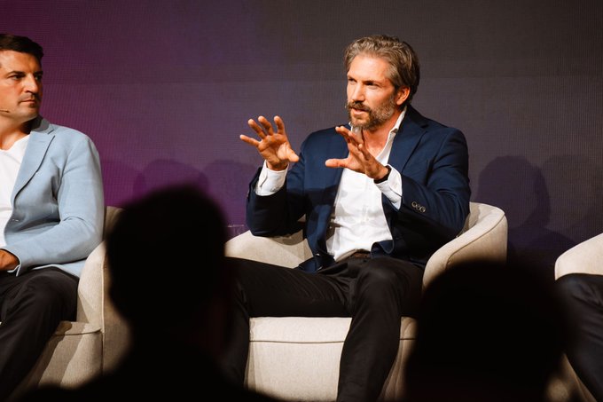 A man with a beard, wearing a navy blazer and white shirt, gestures expressively with both hands while seated on a white chair during what appears to be a panel discussion.