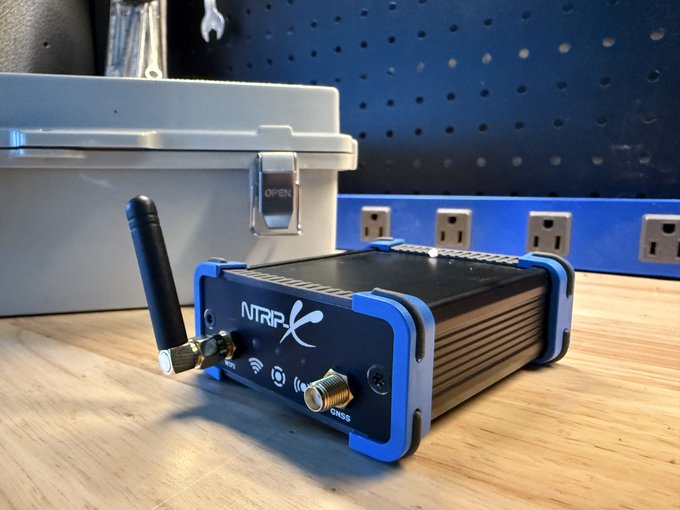A small blue and black electronic device labeled 'NTRIP' with two antennas, sitting on a wooden workbench in front of a blue pegboard with electrical outlets.
