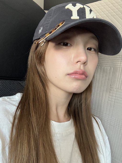 A selfie of a young woman wearing a navy New York Yankees cap with a hair clip, glossy lips, and a white t-shirt.