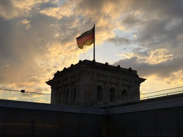 The image features a large flag flying high over a building at sunset. The sky above the building has a beautiful orange hue, creating an impressive backdrop for the flag. The sun is setting behind the building, casting long shadows and highlighting the architectural details of the structure. In the background, there are other buildings visible as well.