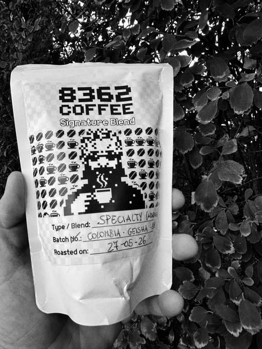 A black and white photo of a hand holding a '8362 COFFEE Signature Blend' bag with a QR code design, labeled as SPECIALTY from Colombia Geisha, roasted on 24-08-26.