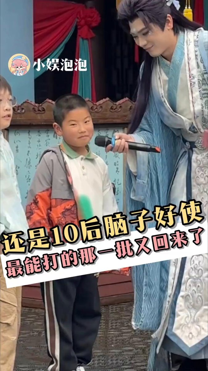 The image features a young boy standing next to two other children, all dressed up for an event or performance. The boy with the microphone appears to be giving instructions or directions to the other children. The background of the photo includes a building, suggesting that they might be in a public place like a park or a community center. The post also contains Chinese text and a picture of a man's head, which could potentially be related to the event taking place or the theme of the performance.