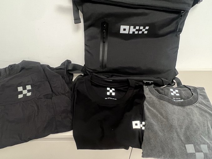 A collection of OKX-branded black and gray apparel and a backpack arranged on a light surface.