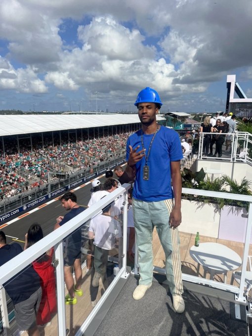 A man wearing a blue hard hat and blue t-shirt posing at what appears to be the Crypto.com Miami Grand Prix venue, with crowds and the racetrack visible in the background.