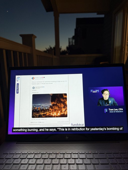 A laptop on an outdoor balcony at dusk (with Venus visible in the sky) plays a Fundstrat "Macro Minute" featuring Tom Lee, CFA, discussing news about a bombing in retribution, as shown on the screen's tweet.
