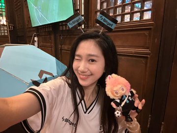 A selfie of a young woman wearing a light-colored jersey, holding a small plush doll and an umbrella in what appears to be an indoor venue with screens in the background.