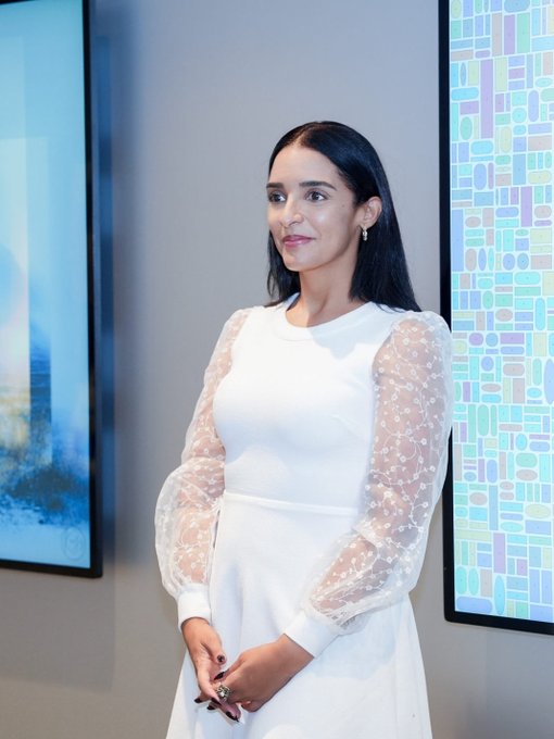 A woman with long dark hair wearing an elegant white dress with sheer floral-embroidered sleeves stands in front of colorful abstract artwork.