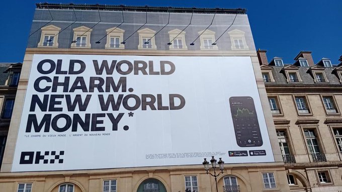A large billboard on a Parisian-style building reads 'OLD WORLD CHARM. NEW WORLD MONEY.' with French translation 'LE CHARME DU VIEUX MONDE. L'ARGENT DU NOUVEAU MONDE' and an OKX ad.