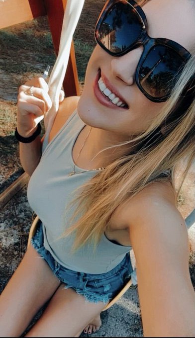 A selfie of a smiling blonde woman wearing dark sunglasses, a light green top, and a delicate cross necklace.