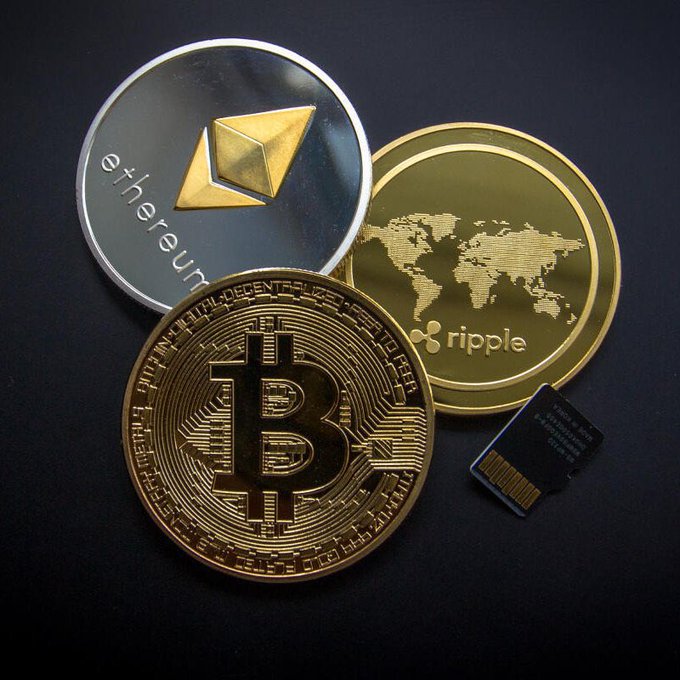 A photograph of physical cryptocurrency coins including Bitcoin, Ethereum, and Ripple (XRP), along with an SD card, arranged on a dark surface.