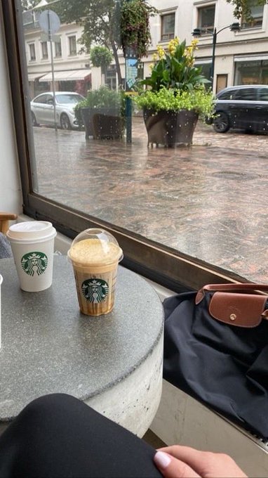 Two Starbucks cups sit on a stone table next to a window overlooking a rainy city street with planters and parked vehicles.