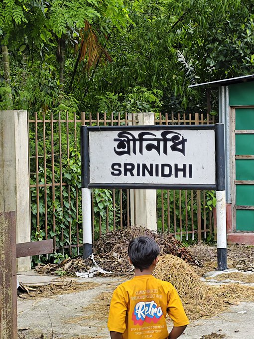 The image features a young boy wearing a yellow shirt standing near a sign that says "Srinidhi" written in an Asian script, possibly Hindi or Bengali. The boy appears to be looking at something off-camera, with his attention seemingly captured by the sign and its surroundings.