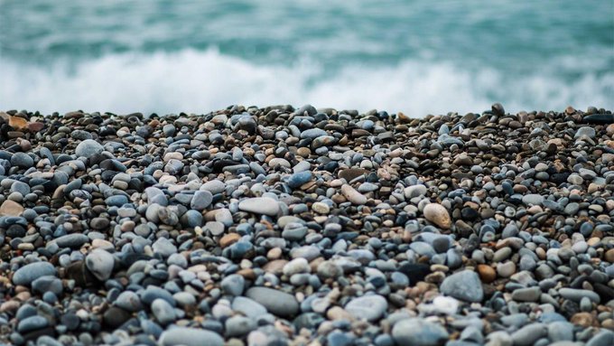 A close-up of smooth, multicolored pebbles on a beach with ocean waves visible in the blurred background.