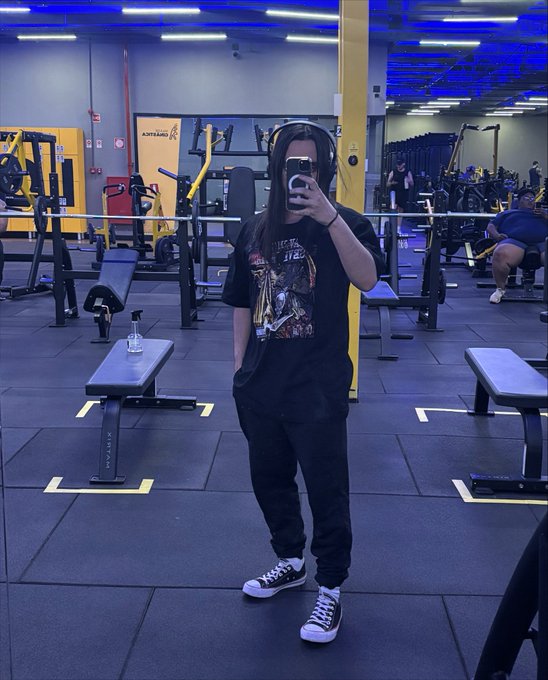 A person taking a mirror selfie in a gym, wearing a black graphic t-shirt and dark jeans, holding a phone that obscures their face.