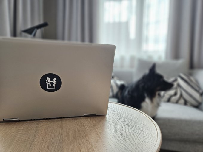 The image shows a laptop computer sitting on top of a wooden table, with a dog resting its head on the table next to it. The background features a couch and curtains, creating an indoor setting for the scene. The caption reads "GM @Eldorim_xyz Loading".