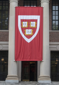 A large crimson banner bearing the Harvard University shield with the word "VERITAS" hangs between classical stone columns of a campus building. The image visually anchors the post's news about Harvard's updated ETF holdings, with the prestigious institution's emblem symbolizing the source behind its reported $101.4 million BlackRock IBIT position and $171.2 million gold holdings.
