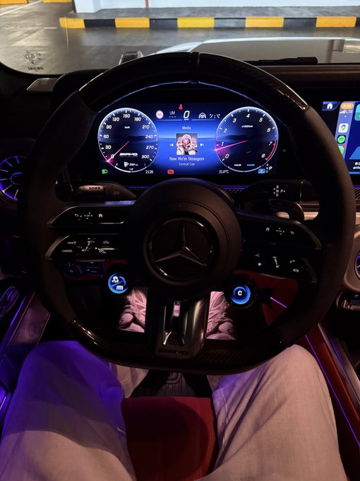 A driver sits behind the wheel of a Mercedes-Benz at night in what appears to be a parking garage, with the dashboard display showing "Now We're Strangers" by Arma Cor. The "POV you bought BTC and HYPE at the bottom" caption playfully contrasts the luxurious, relaxed moment behind the wheel with the risky crypto investment, suggesting the driver is in the calm before either a surge or a steep drop.