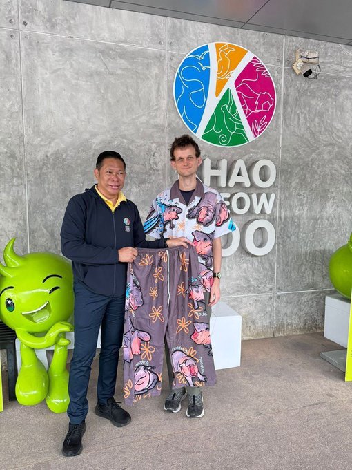 Two men posing together in front of a wall with a colorful animal logo and partial text 'HAO' and 'DO'. One wears a dark jacket, while the other is dressed in a purple kimono-style garment with pink patterns, standing next to a green cartoon mascot statue.