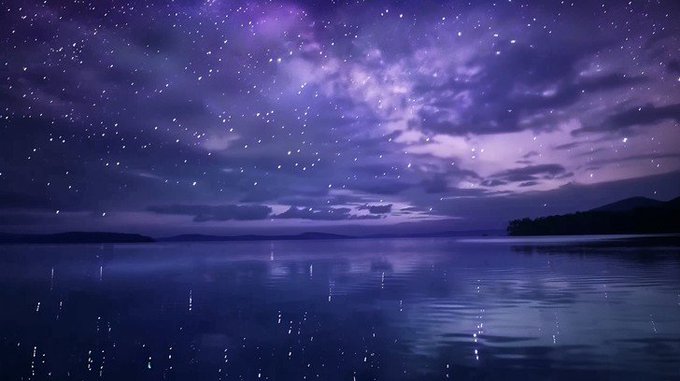 A serene nighttime landscape showing a starry sky with the Milky Way visible in purple and blue hues, reflected on the calm surface of a lake with distant mountains on the horizon.