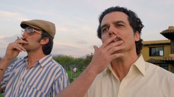 Two men, one wearing a tan cap and sunglasses and the other with curly hair, both smoke cigarettes while looking upward in an outdoor setting with a building and landscape in the background.
