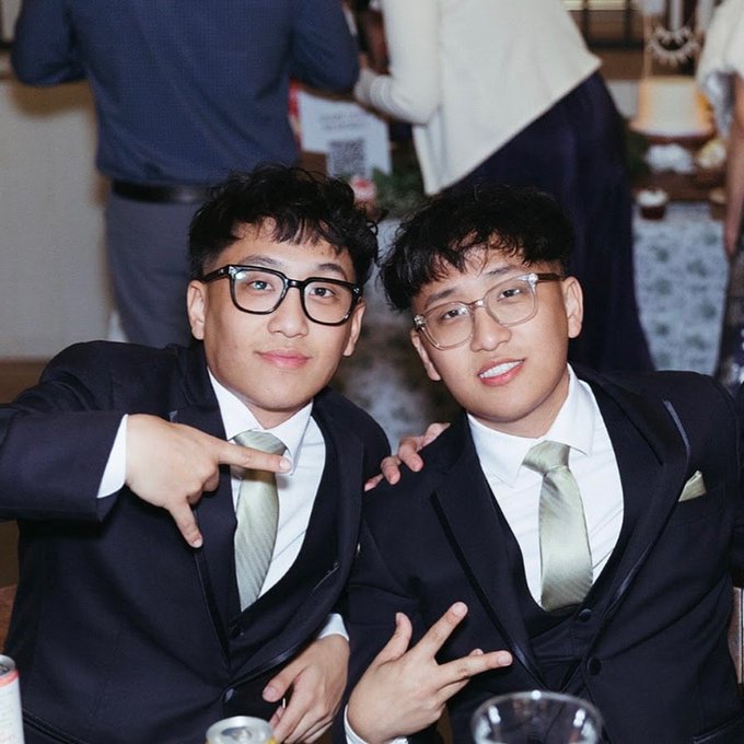 Two well-dressed individuals in matching black suits, white shirts, and sage green ties pose for the camera, flashing peace signs with friendly smiles and glasses. The image captures a social moment that aligns with the post's caption about the "degentwinzz" — likely featuring the duo at a casual gathering where trading tips are being shared.