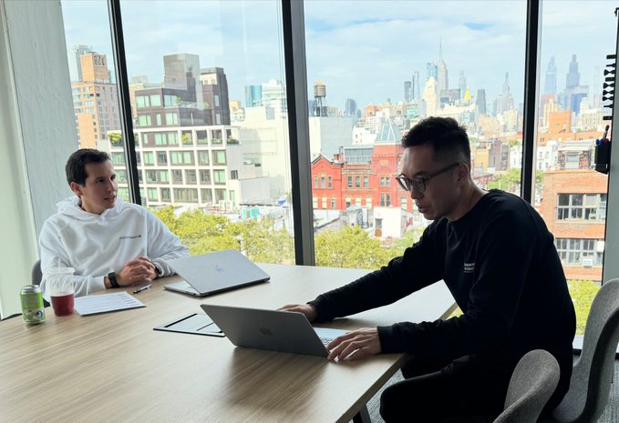 Two men work on laptops at a wooden table in a modern office with large floor-to-ceiling windows overlooking a city skyline with skyscrapers.