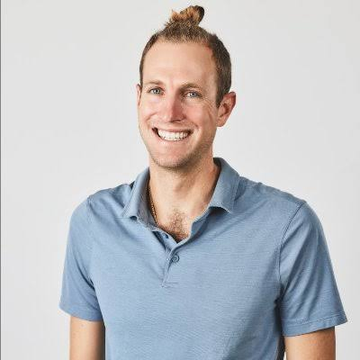 A smiling man with a man bun wearing a blue polo shirt poses for a portrait against a light gray backdrop.
