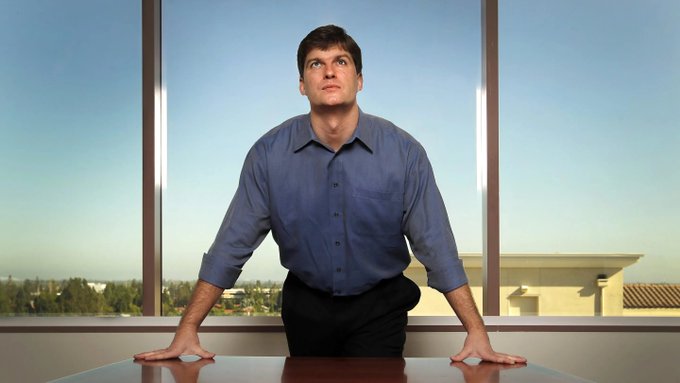 A man in a blue dress shirt leans forward on a conference table, staring upward with a concerned, dazed expression, with large windows showing a suburban landscape behind him—his worried look visually reinforcing the post's skeptical commentary about his poor track record and habitual short positions since 2008.